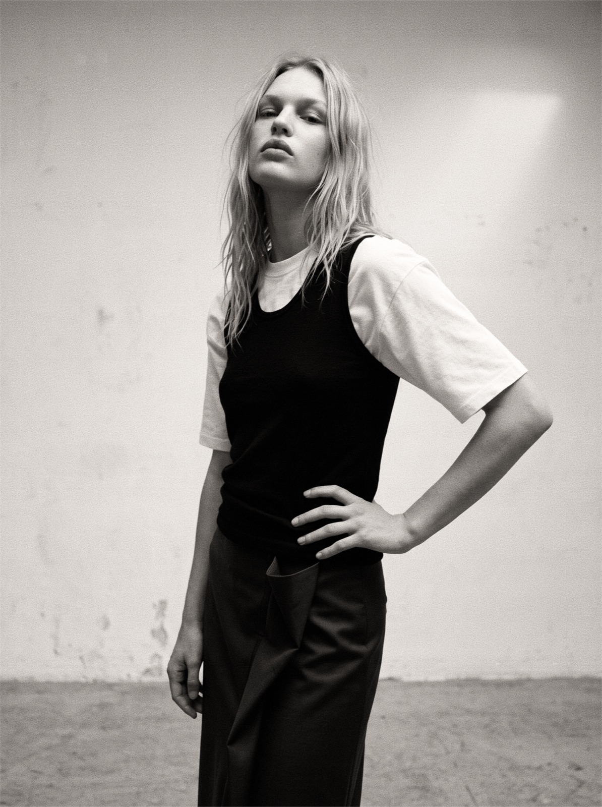 Image — a lookbook portrait against a raw concrete wall, hands on hips