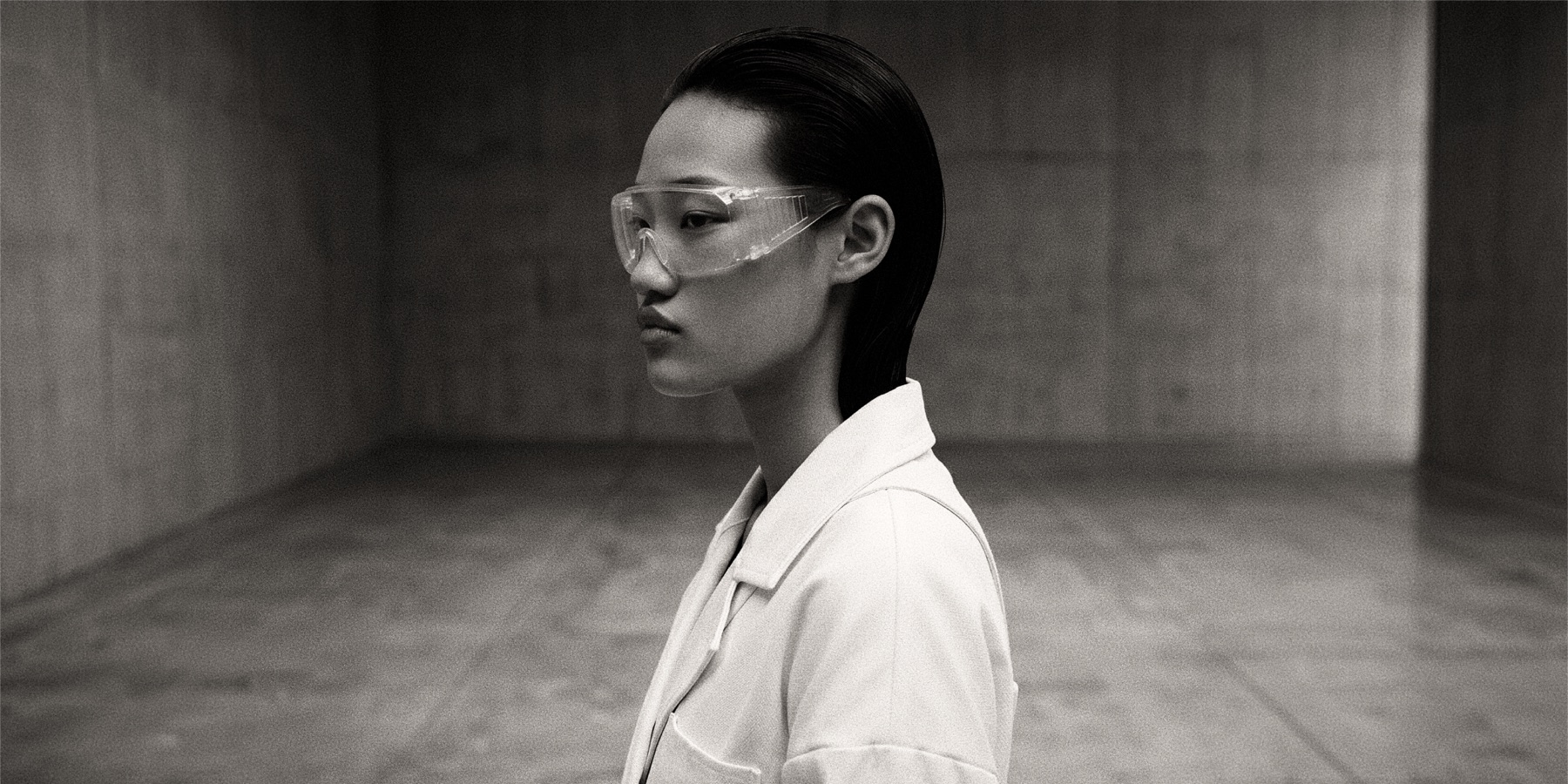 Re-Authored — a profile portrait in clear lab goggles and a white coat, a concrete room behind her