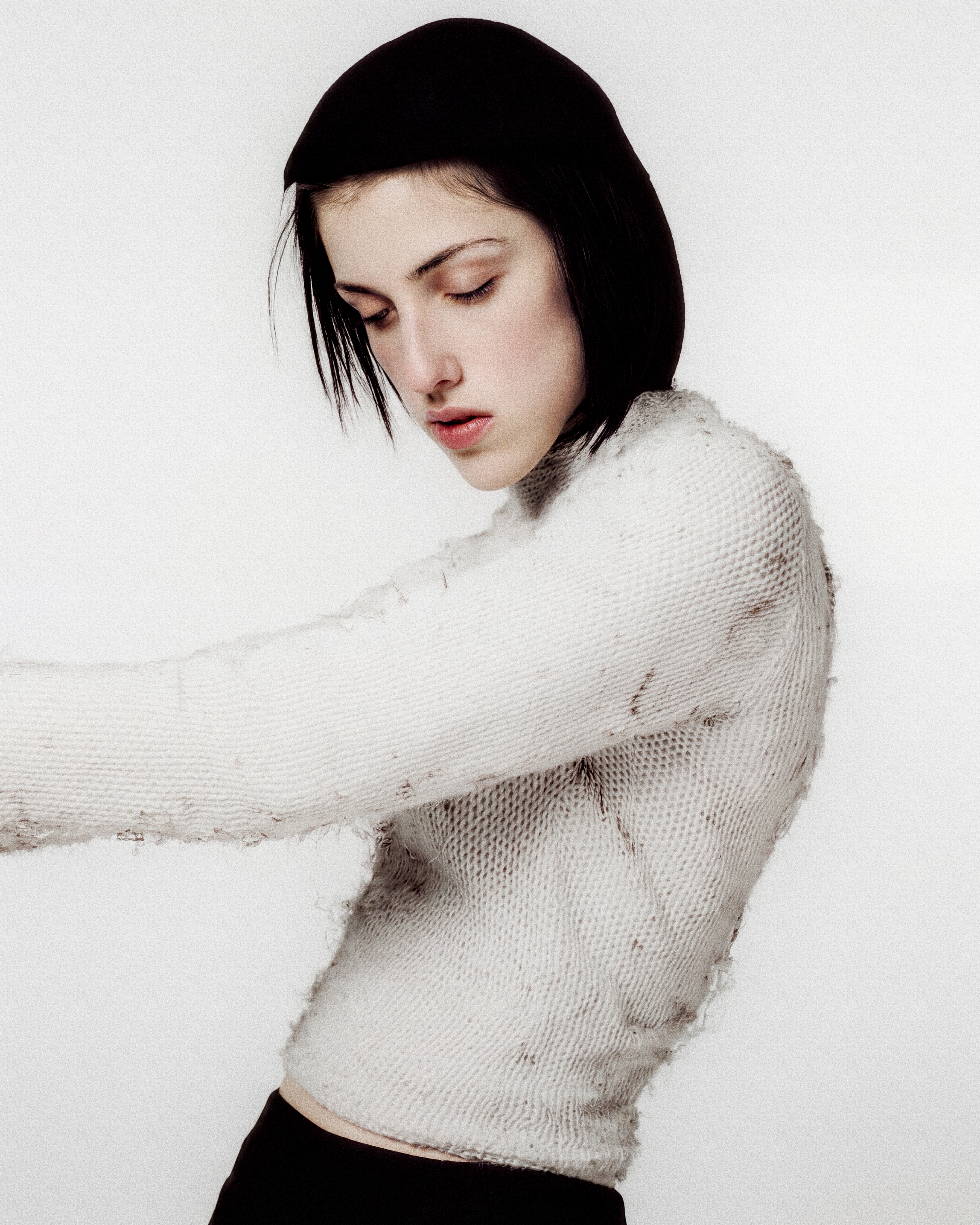 Portrait of a dark-haired figure in a distressed white knit, leaning forward against a white field.